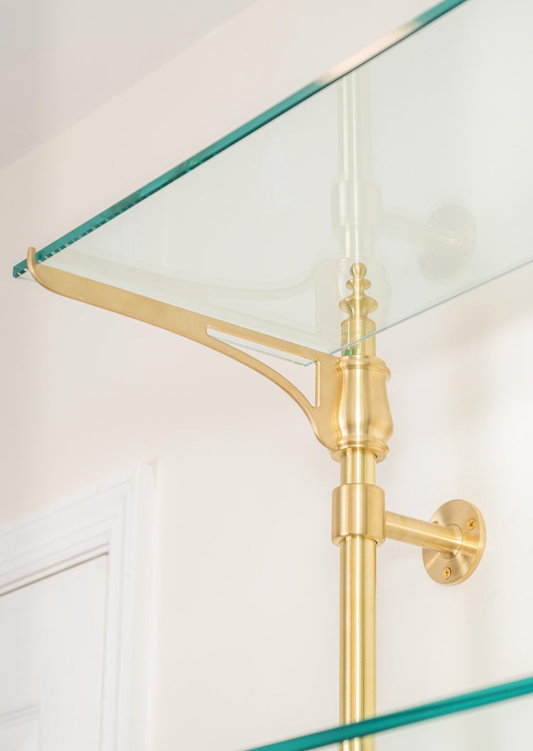 An ornate cantilever arm of a bistro shelving unit made from real brass that is supporting a glass shelf.
